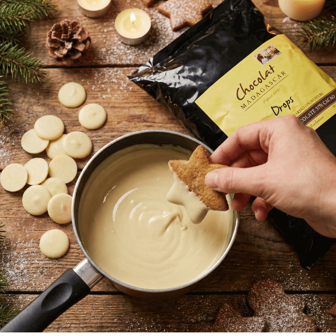Top-down view of a festive Christmas baking scene featuring a hand dipping a star-shaped cookie into a saucepan of melted white chocolate. The rustic wooden table is arranged with scattered white chocolate drops, a large bag of Chocolat Madagascar White Gold couverture, pine branches, red berries, and lit candles.
